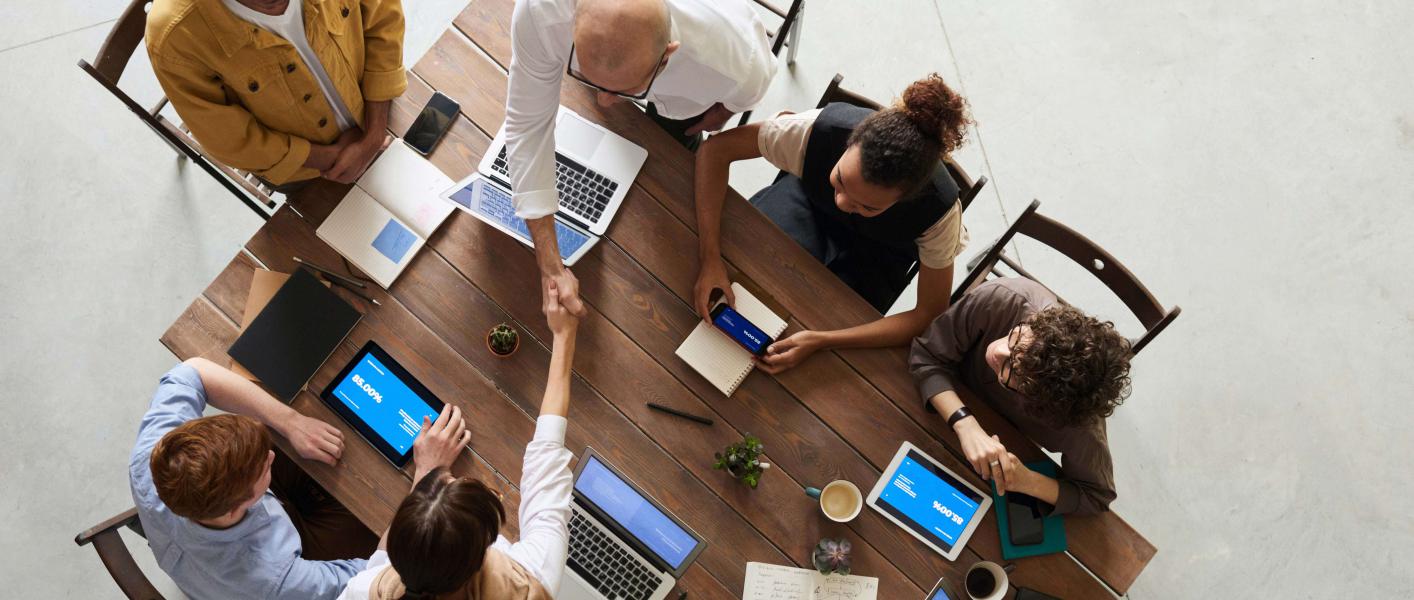 seis personas reunidas en una mesa, con equipos portátiles y celulares, charlando. dos personas se dan la mano