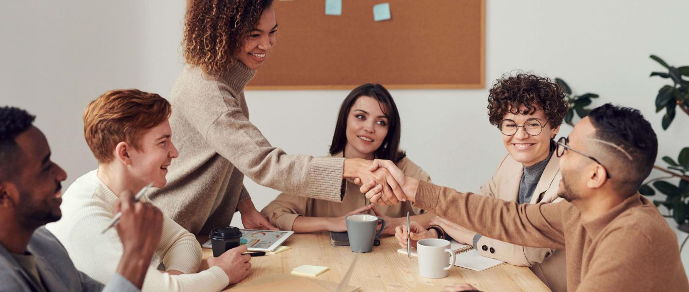 6 personas reunidas, en una mesa. Una persona de pié le da la mano a otra que está sentada